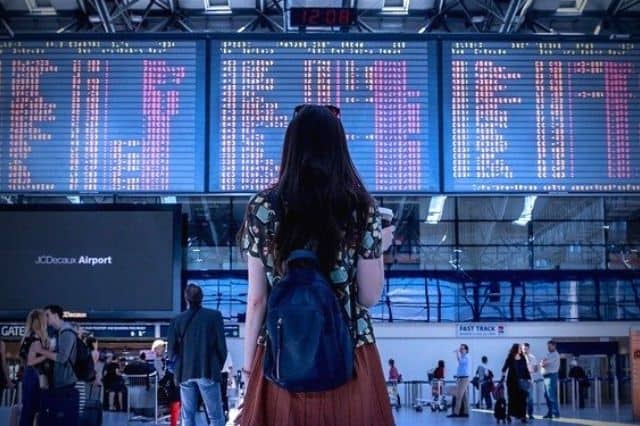 A young girl is facing away from the camera looking at a big board of departing flights in a crowded airport