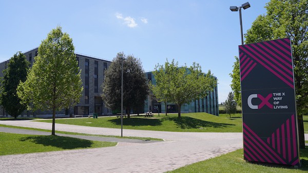 A picture overlooking some of the buildings in Campus X, there is plenty of greenery and some trees, with tall square buildings in the back