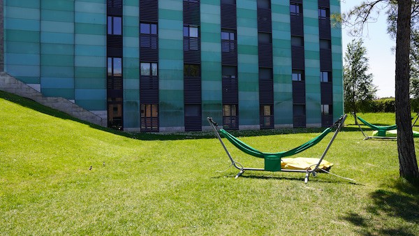 An outdoor hangout spot in cx place, we can see a green hammock on an open grass field with a large and modern square building behind which is the private dorms of Rome.