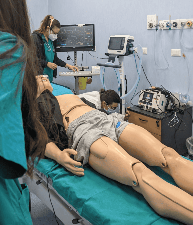 There is a simulation medical dummy lying on a hospita bed with cables hooked up to him and monitors in the background. A doctor is checking an ekg monitor behind him, and another student is looking at the dummy.