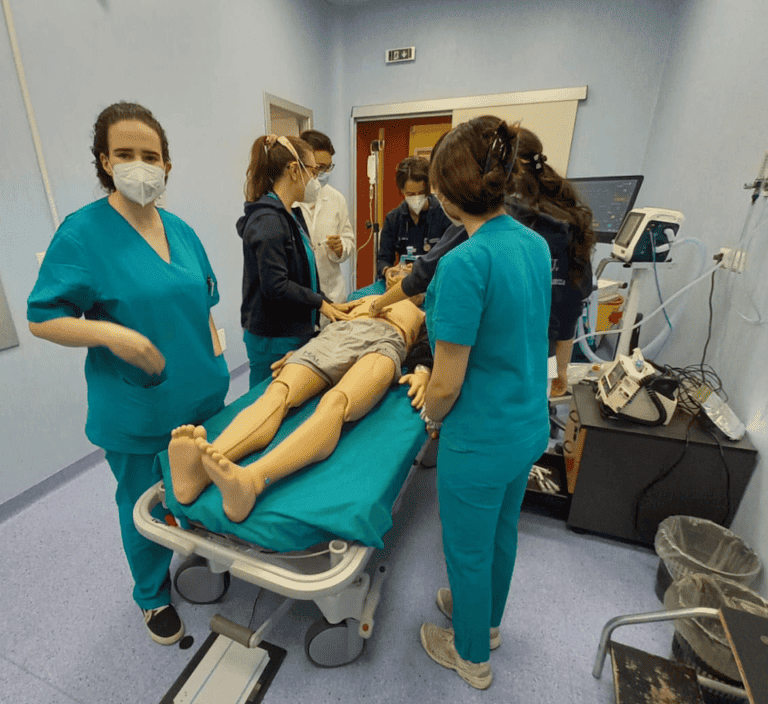 A group of medical students and doctors stand around a table that has a medical simulation dummy on it. There are many monitors and heart rate cables.